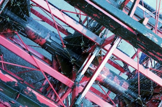 A close-up view of a mechanical structure composed of red and black metal beams and rods, forming a complex, geometric pattern with water splashing against it.