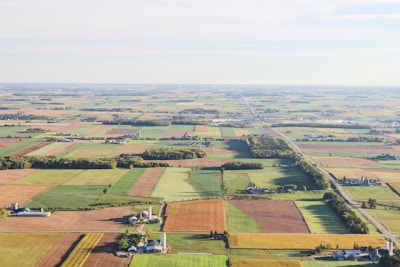 Aerial view of a vast expanse of farmland divided into neatly organized rectangular plots. The fields display a variety of colors, indicating different crops and stages of growth. Scattered farm buildings, silos, and narrow country roads are interspersed throughout the landscape. The horizon reveals a subtle curvature of the earth under a bright, clear sky.