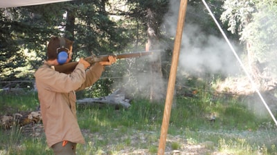 A person is aiming a shotgun outdoors in a wooded area, with smoke visible around the barrel. The shooter is wearing ear protection and a beige jacket. The background is filled with trees and greenery.