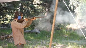 A person is aiming a shotgun outdoors in a wooded area, with smoke visible around the barrel. The shooter is wearing ear protection and a beige jacket. The background is filled with trees and greenery.