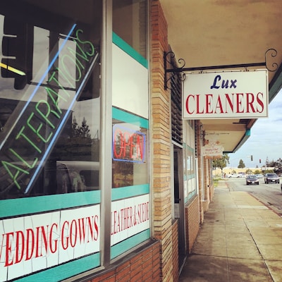 A corner shop features multiple signs including 'Lux Cleaners' in bold red and blue letters. The window displays neon signs for 'Alterations' and 'Open', and other information about services like 'Wedding Gowns' and 'Leather & Suede'. The building has a brick facade with turquoise and white accents. The surrounding area includes a sidewalk and a street with some visible vehicles under a light blue sky.
