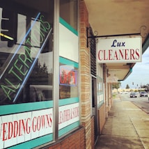 A corner shop features multiple signs including 'Lux Cleaners' in bold red and blue letters. The window displays neon signs for 'Alterations' and 'Open', and other information about services like 'Wedding Gowns' and 'Leather & Suede'. The building has a brick facade with turquoise and white accents. The surrounding area includes a sidewalk and a street with some visible vehicles under a light blue sky.