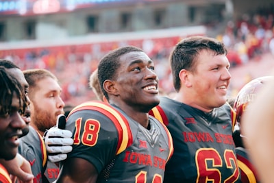A group of young American football players, wearing matching Iowa State uniforms, are standing closely together in what appears to be a celebratory or spirited moment. Their expressions are joyful and they seem to be engaged in camaraderie, likely after a game or significant event. The background shows a stadium filled with fans, though it's out of focus, suggesting a lively atmosphere.