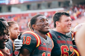 A group of young American football players, wearing matching Iowa State uniforms, are standing closely together in what appears to be a celebratory or spirited moment. Their expressions are joyful and they seem to be engaged in camaraderie, likely after a game or significant event. The background shows a stadium filled with fans, though it's out of focus, suggesting a lively atmosphere.
