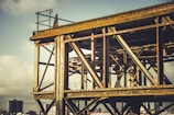 A partially constructed steel structure with a lattice of rusty beams and girders against a cloudy sky. In the background, there are buildings and a cityscape visible, creating an industrial setting.