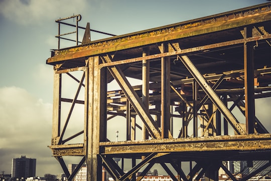 A partially constructed steel structure with a lattice of rusty beams and girders against a cloudy sky. In the background, there are buildings and a cityscape visible, creating an industrial setting.