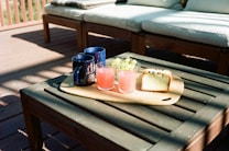 A wooden outdoor coffee table with a light wood serving board holding two glasses filled with pink lemonade, two dark blue mugs, a cluster of green grapes in a glass bowl, and a slice of cake. The table is set on a wooden deck with sunlight casting soft shadows. Cushioned outdoor furniture is positioned in the background.