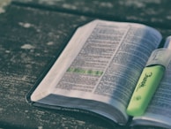 An open book with text and a highlighted line in green, resting on a textured surface. A green highlighter is placed on the book.