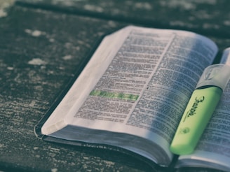An open book with text and a highlighted line in green, resting on a textured surface. A green highlighter is placed on the book.