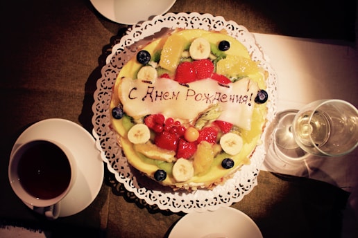 A colorful fruit cake with slices of kiwi, banana, orange, and strawberries is adorned with a message written in Cyrillic script, placed on a delicate lace doily. Nearby, there's a cup of tea and a glass of wine on a table.