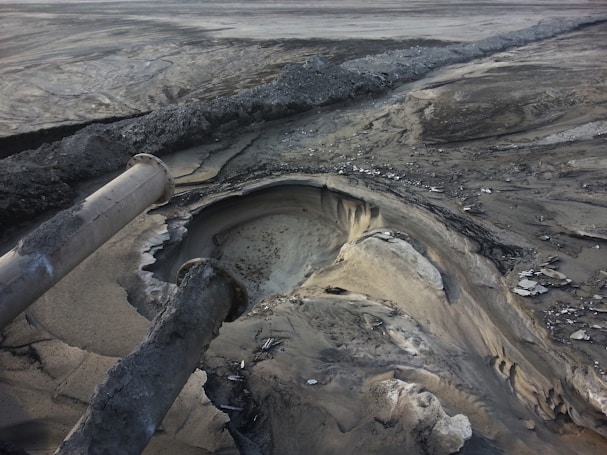 Two large industrial pipes protrude over a barren, desolate landscape, leading to a deep crater-like pit surrounded by layers of sediment. Sparse, cracked earth stretches into the distance, hinting at a site of extensive excavation or environmental disturbance.