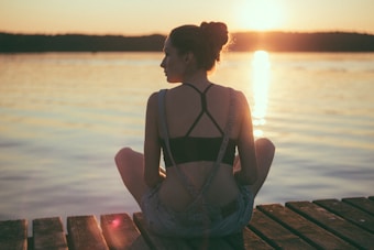 A person is sitting cross-legged on a wooden dock gazing at the sunset over a calm lake. The warm glow of the setting sun creates a peaceful and reflective atmosphere.