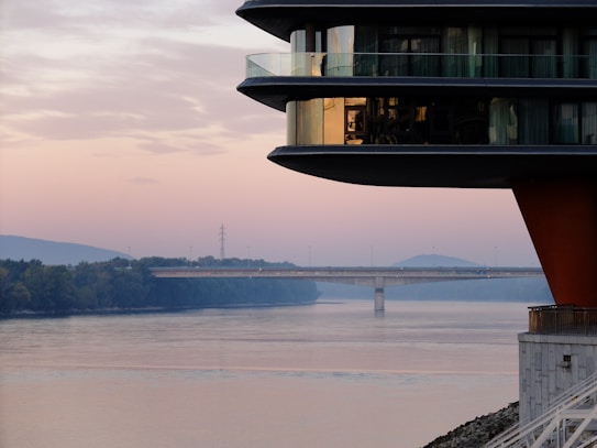 A modern building with glass balconies is prominently featured on the right side, overlooking a calm river with a bridge in the distance. The sky displays a soft gradient of pink and blue as the sun sets or rises. The surroundings include lush greenery and distant hills, adding to the serene atmosphere.