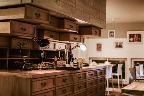 A cozy kitchen with wooden cabinets, multiple drawers, and shelves. There is ambient lighting created by two desk lamps on the countertop. The counter is organized with various objects, including a wicker basket and small potted plant. The background includes a dining area with chairs and framed pictures hanging on the wall.