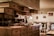 A cozy kitchen with wooden cabinets, multiple drawers, and shelves. There is ambient lighting created by two desk lamps on the countertop. The counter is organized with various objects, including a wicker basket and small potted plant. The background includes a dining area with chairs and framed pictures hanging on the wall.