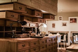 A cozy kitchen with wooden cabinets, multiple drawers, and shelves. There is ambient lighting created by two desk lamps on the countertop. The counter is organized with various objects, including a wicker basket and small potted plant. The background includes a dining area with chairs and framed pictures hanging on the wall.