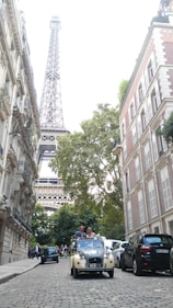 A narrow cobblestone street is flanked by classic European buildings with ornate architecture. In the background, the Eiffel Tower looms against the sky. A vintage cream car with a couple, possibly newlyweds, stands out on the street. Around them, a few other cars and pedestrians can be seen.