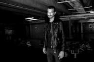 A model wearing a minimalist tribal leather coat, standing in a shadowed urban setting with cinematic contrast.