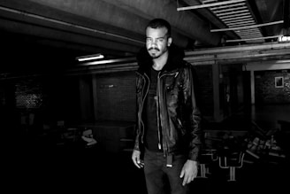A person stands in a dimly lit urban setting, wearing a dark leather jacket with a fur collar. The environment has an industrial feel, with exposed metal beams and brick walls. The lighting creates dramatic contrasts, emphasizing the subject's features against the shadowy background.