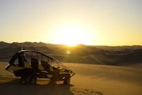 A dune buggy is parked on a sandy desert landscape with vast, smooth dunes stretching into the distance. The sun is setting on the horizon, casting a warm, golden light over the scene.