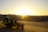 A dune buggy is parked on a sandy desert landscape with vast, smooth dunes stretching into the distance. The sun is setting on the horizon, casting a warm, golden light over the scene.