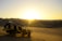 A dune buggy is parked on a sandy desert landscape with vast, smooth dunes stretching into the distance. The sun is setting on the horizon, casting a warm, golden light over the scene.