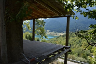 A partially constructed wooden platform extends from a structure, surrounded by lush green trees. In the distance, a tranquil lake is nestled among forested hills and mountains under a clear blue sky.