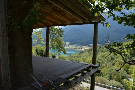 A partially constructed wooden platform extends from a structure, surrounded by lush green trees. In the distance, a tranquil lake is nestled among forested hills and mountains under a clear blue sky.