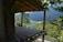 A partially constructed wooden platform extends from a structure, surrounded by lush green trees. In the distance, a tranquil lake is nestled among forested hills and mountains under a clear blue sky.