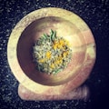 A marble mortar and pestle containing a mixture of herbs and spices, including dried rosemary and yellow turmeric powder, placed on a dark speckled countertop.