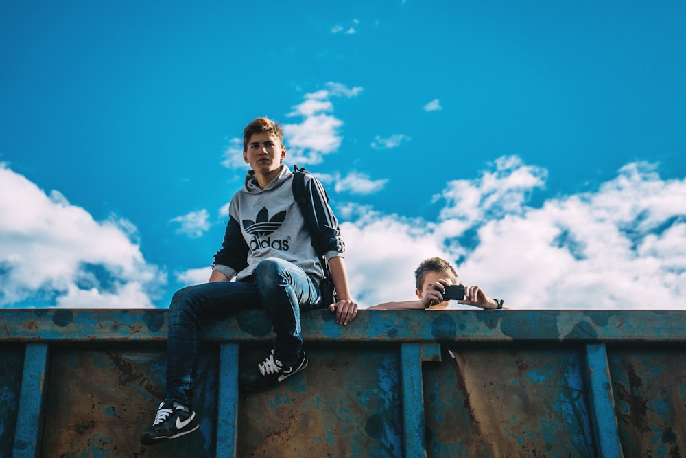 man sitting on rail beside man holding camera