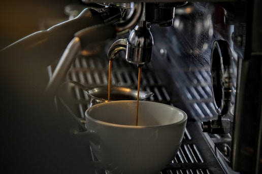 Espresso coffee pours into a white ceramic cup from a metal espresso machine spout, creating a rich and inviting scene. The focus is on the dark coffee flowing smoothly, surrounded by a cozy, dimly lit atmosphere that highlights the stainless steel and shiny surfaces of the machine.