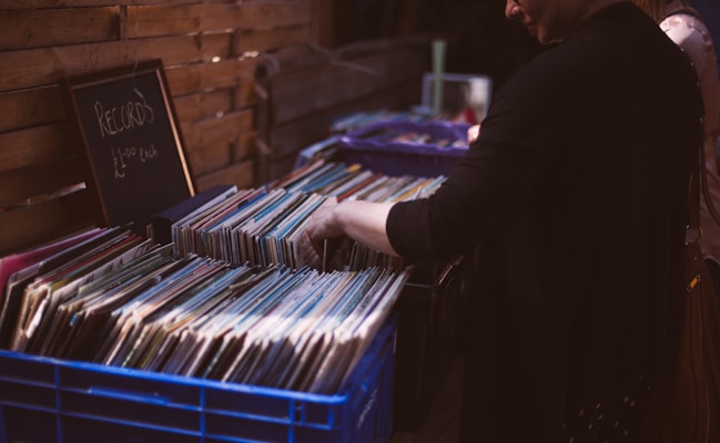 A person browses through a collection of vinyl records stored in blue plastic crates. A chalkboard sign next to the crates reads 'Records &pound;1.00 each'. The setting appears to be an outdoor market or shop with wooden walls visible in the background.