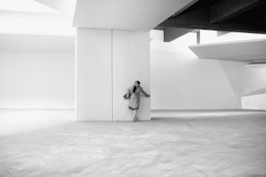 A person in a coat stands against a large, white architectural structure in a minimalist setting. The design includes strong geometric lines and shadows, emphasizing the contrast between light and dark. The setting is spacious and devoid of additional objects, contributing to a sense of simplicity and elegance.