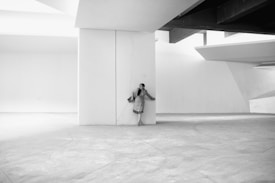 A person in a coat stands against a large, white architectural structure in a minimalist setting. The design includes strong geometric lines and shadows, emphasizing the contrast between light and dark. The setting is spacious and devoid of additional objects, contributing to a sense of simplicity and elegance.