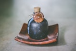 A dark-colored, round bottle with a cork stopper labeled 'Body Lotion' is placed on a wooden dish. The bottle has a rustic and vintage appearance, set against a soft, blurred background.