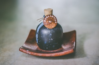 A dark-colored, round bottle with a cork stopper labeled 'Body Lotion' is placed on a wooden dish. The bottle has a rustic and vintage appearance, set against a soft, blurred background.