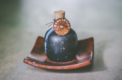 A dark-colored, round bottle with a cork stopper labeled 'Body Lotion' is placed on a wooden dish. The bottle has a rustic and vintage appearance, set against a soft, blurred background.