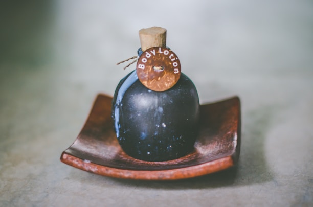 A dark-colored, round bottle with a cork stopper labeled 'Body Lotion' is placed on a wooden dish. The bottle has a rustic and vintage appearance, set against a soft, blurred background.