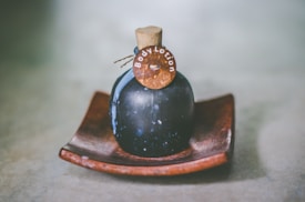 A dark-colored, round bottle with a cork stopper labeled 'Body Lotion' is placed on a wooden dish. The bottle has a rustic and vintage appearance, set against a soft, blurred background.