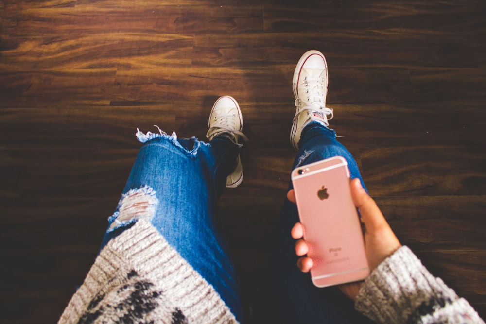 Person mit roségoldenem iPhone 6s in einer blauen Jeans im Used-Look, die auf braunem Holzboden steht