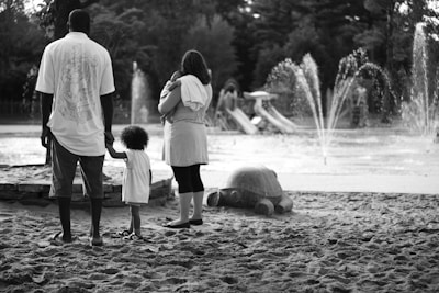A black and white image of a family at a park, with an adult man holding a child's hand and a woman carrying an infant standing nearby. The scene includes a sandy area, a large turtle sculpture, and water fountains with spray in the background. Trees and playground slides are visible in the distance.