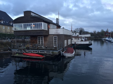 A wooden houseboat with large windows is moored on a calm body of water. A red kayak and a small white boat are tied up to the side of the houseboat. Another larger boat is moored nearby. The surroundings include other buildings and some trees, under a cloudy sky.