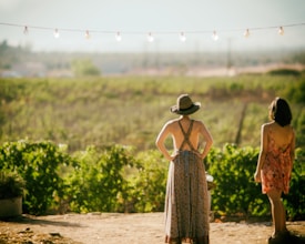 Two people wearing summer dresses standing outdoors, facing away towards a vast, open field. One person is wearing a wide-brimmed hat. There are string lights hanging above them, and lush green foliage surrounds the area.