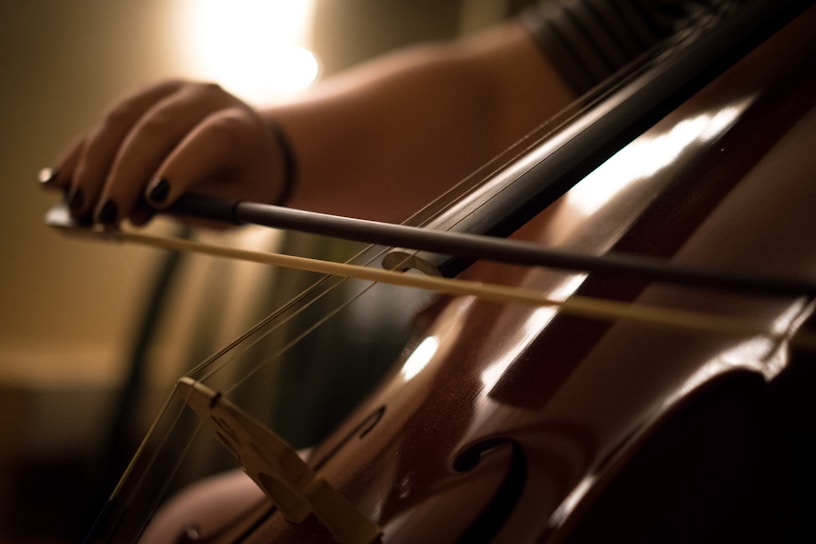 A close-up view of a person's hand playing a cello with a bow, highlighting the strings and wooden body of the instrument. The lighting creates a warm and intimate atmosphere, with a focus on the details of the hand and the cello.