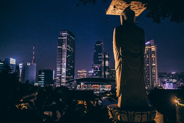 A large statue of Buddha is silhouetted against the backdrop of a city skyline at night. The buildings are illuminated with lights, showcasing various architectural designs. The scene conveys a blend of spirituality and urban life.
