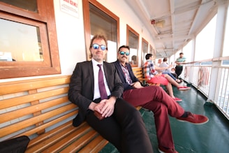 Two men in formal attire and sunglasses sit on a wooden bench on a large deck, with several other casually dressed people in the background. The deck has green flooring and white railings, and there's a row of wooden framed windows on the side.