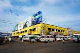 A busy street scene featuring a large yellow building with multiple billboards on the roof, showcasing advertisements. Several vehicles, including buses and vans, are parked and moving around. Pedestrians can be seen walking along the street, with some standing near the vehicles. Overhead wires and blue decorative flags are also visible.