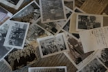 A collection of vintage black and white photographs scattered over a surface, some containing groups of people posing outdoors and others featuring formal portraits. Handwritten notes are visible on some photographs, suggesting dates and names.