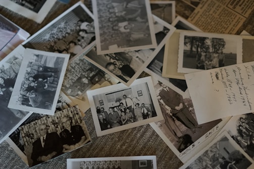 A collection of vintage black and white photographs scattered over a surface, some containing groups of people posing outdoors and others featuring formal portraits. Handwritten notes are visible on some photographs, suggesting dates and names.